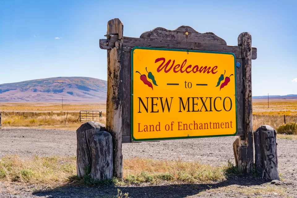"Welcome to New Mexico - Land of Enchantment" roadside sign with red text onyellow background and rustic wood frame around sign.